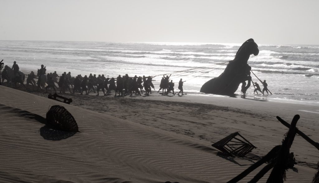 The Trojan Horse hauled from the surf up a beach with ropes by a long line of figures — Nolan's Odyssey (2026).